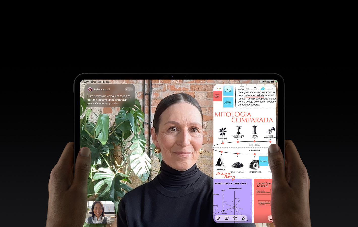 Parte da frente do iPad Pro preto-espacial na horizontal, mostrando os cantos arredondados e a tela com borda preta. Duas mãos seguram os lados esquerdo e direito. A tela mostra uma conversa no FaceTime sendo traduzida ao vivo. À direita, um app educativo mostra uma apresentação de Contos Épicos com textos, diagramas e notas manuscritas