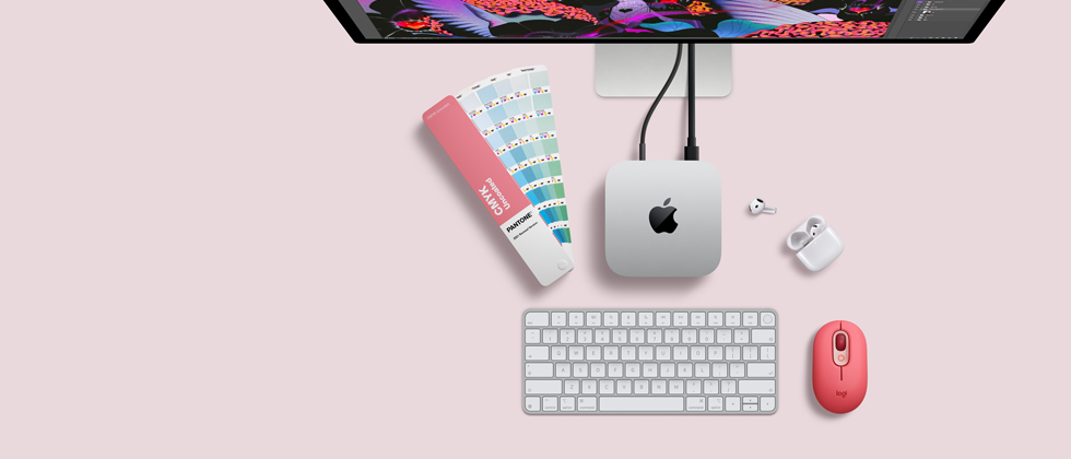 „Mac mini“ iš viršaus: sidabro spalvos aliuminio korpusas, užapvalinti kampai, centre – blizgus juodas „Apple“ logotipas, prie įrenginio prijungtas laidas; viršuje iš dalies matomas „Studio Display“, atidarytas „AirPods 4“ dėklas, „Pantone“ spalvų pavyzdžiai, skaičių klaviatūra ir pelė