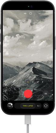 iPhone 18 Pro, priekšpuses skats, redzama kalnu ainava ar kameras režīma pārklājumu Time-Lapse režīmā, strāvas kabelis pievienots uzlādes portam