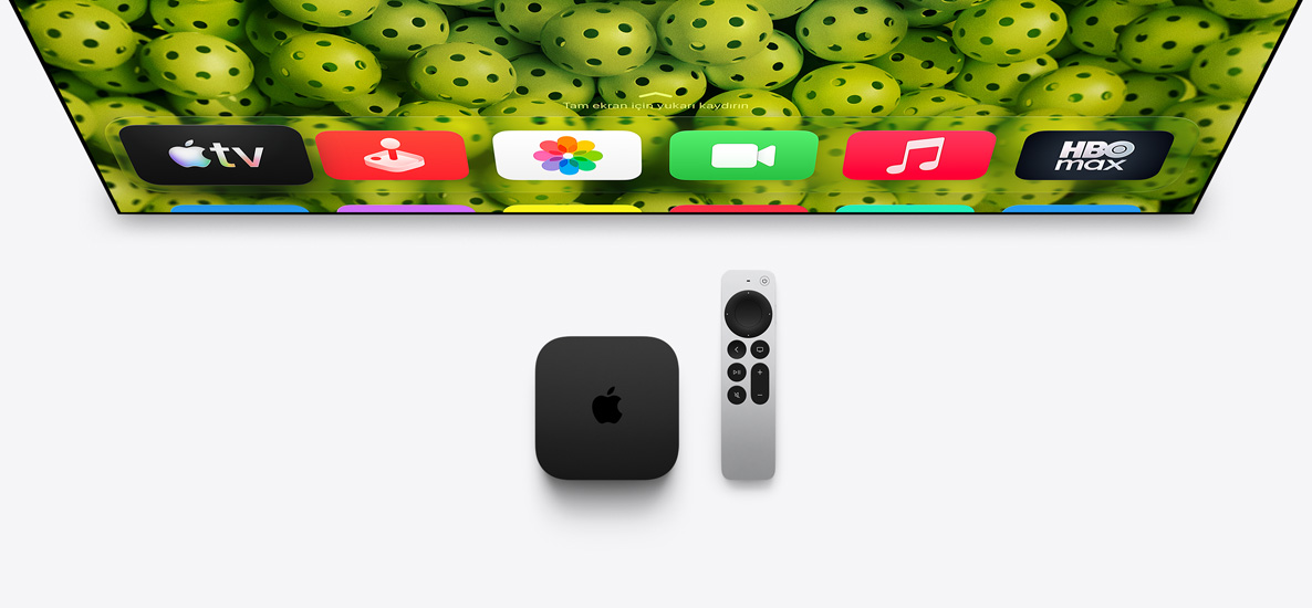 TV ekranının altında Apple TV 4K ve Siri Remote duruyor, ekranda pickleball toplarından oluşan arka plan ve Ana Ekran’daki uygulama simgelerinin üst satırı gösteriliyor