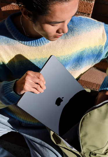 A student sitting outside. He pulls a closed MacBook from his backpack.