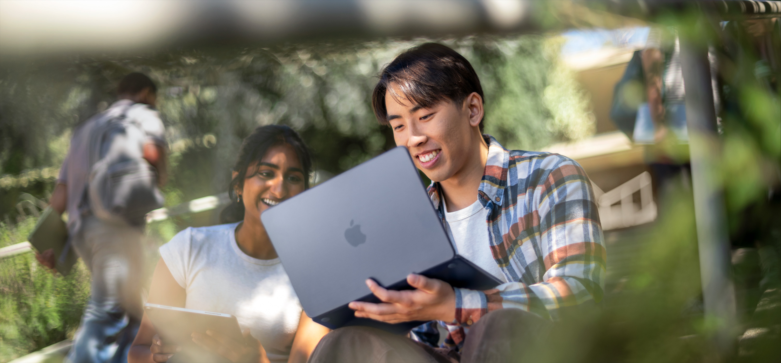 Kaksi hymyilevää opiskelijaa käyttää MacBookia ulkona. Toinen pitää sitä sylissään heidän työskennellessään yhdessä tehtävän parissa