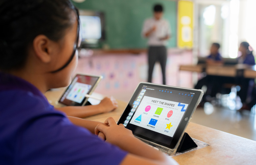 特寫畫面中，一名學生坐在課桌前使用 iPad。螢幕上顯示著色彩繽紛的形狀，標題為 Meet the shapes (認識形狀)。