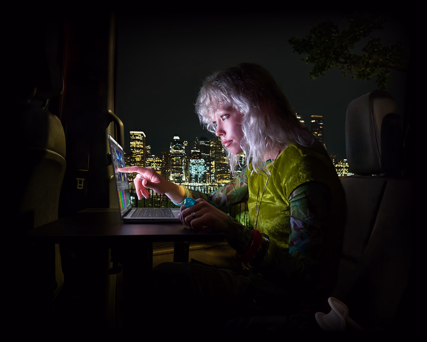 En person sidder på bagsædet af en minibus, den åbne passagerdør viser en naturskøn by ved nattetide, MacBook Pro med PROLED XDR-skærm på et skrivebord, personen oplyses af skærmen, mens vedkommende bruger touch-funktionen til at redigere en video i DaVinci Resolve Studio
