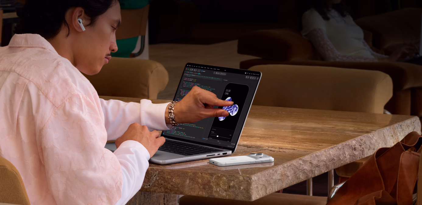 En person sidder ved et bord et offentligt sted og bruger et knibebevægelse til at justere et element i Xcode ved hjælp af touch-funktionen på en sølvfarvet MacBook Pro med PROLED XDR-skærm. En iPhone ligger på bordet ved siden af computeren, og personen bruger AirPods Pro